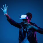 Portrait of young african-american woman's playing in VR-glasses in neon light on blue background. Concept of human emotions, facial expression, modern gadgets and technologies. Climbing up in gameplay.
